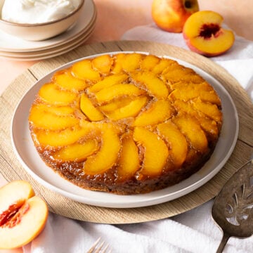 A round peach upside-down cake with glazed peach slices on top, placed on a white plate, surrounded by fresh peaches and a bowl of whipped cream.