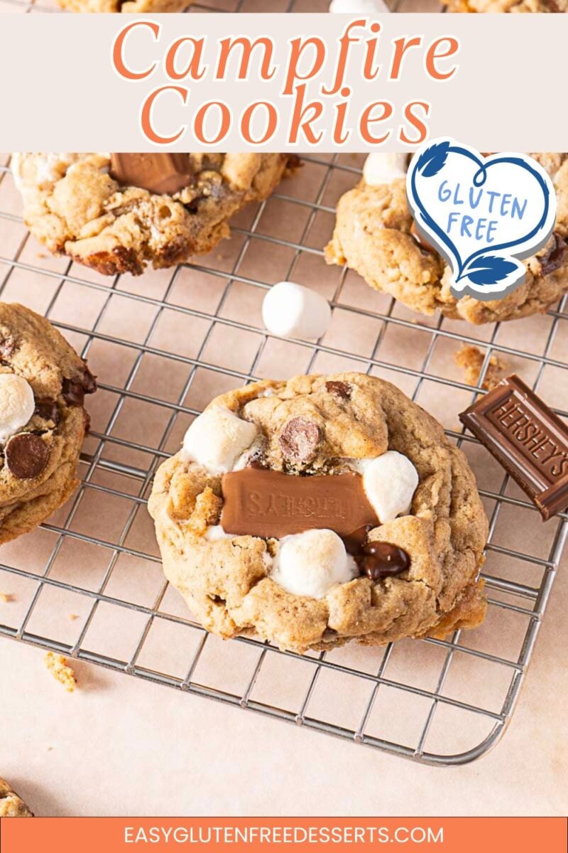 Three S’Mores Cookies topped with marshmallows, chocolate chunks, and chocolate squares cool on a wire rack. A “Gluten Free” symbol and “Campfire Cookies” text appear at the top, highlighting these irresistible Gluten Free Cookies.