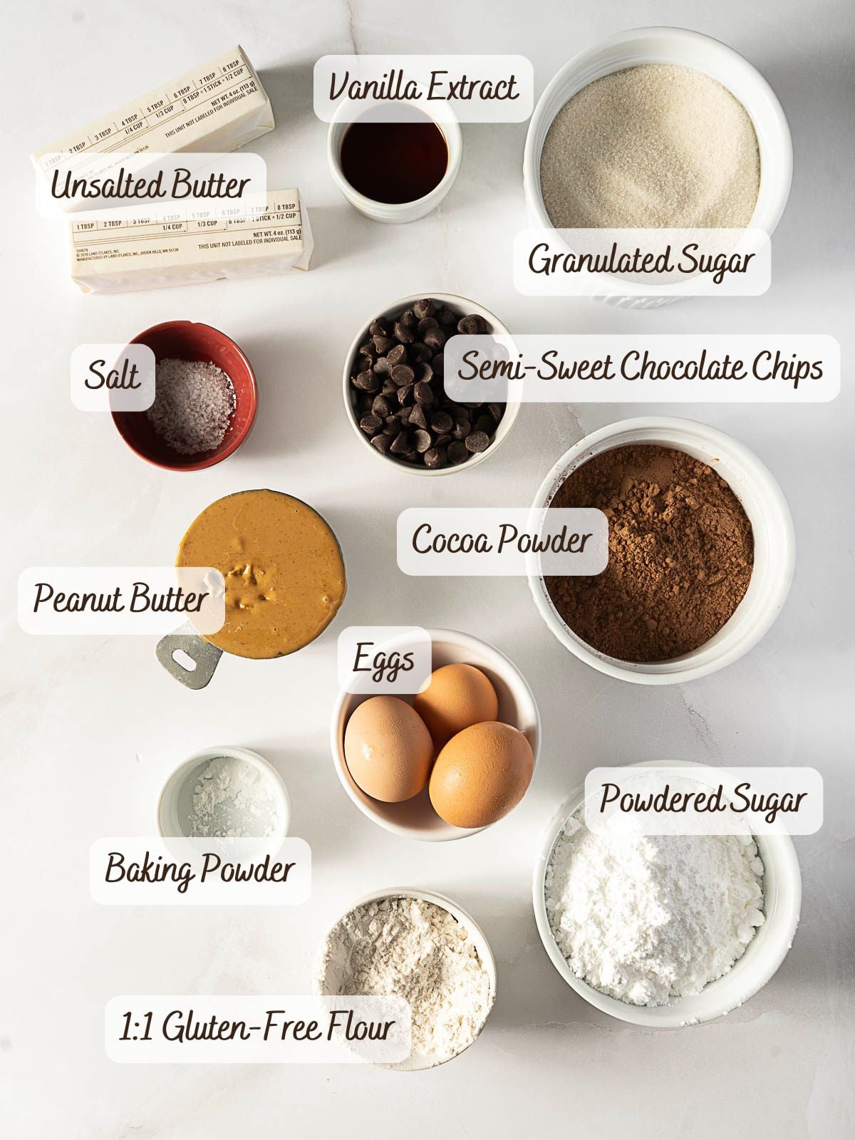 Baking ingredients arranged on a white surface, including eggs, peanut butter, butter, sugar, powdered sugar, flour, cocoa, chocolate chips, vanilla extract, salt, and baking powder