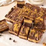 Sixteen square brownies with a marbled chocolate and peanut butter topping are arranged on parchment paper, with a few pieces separated from the main group.
