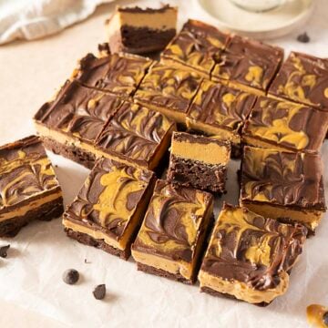 Sixteen square brownies with a marbled chocolate and peanut butter topping are arranged on parchment paper, with a few pieces separated from the main group.