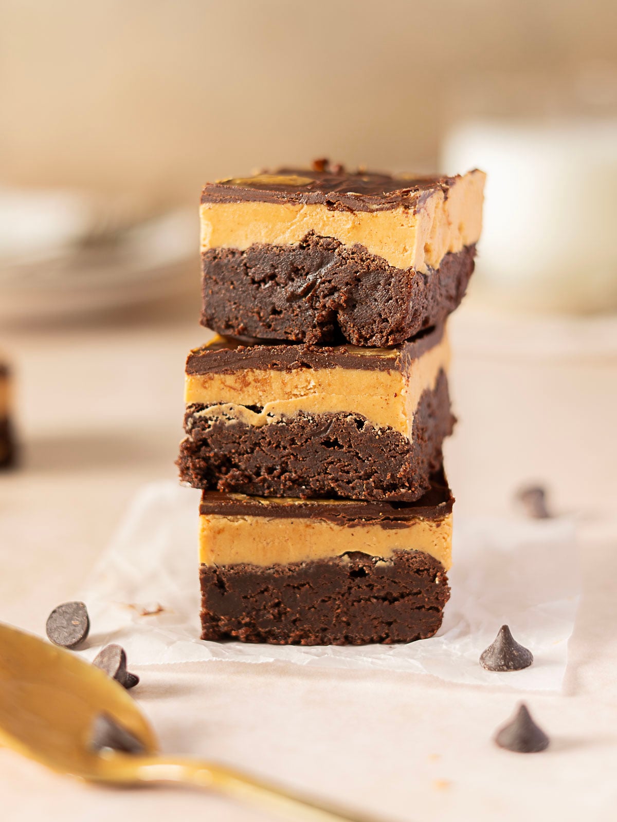 Three chocolate brownies with a thick peanut butter layer are stacked on parchment paper, surrounded by chocolate chips.