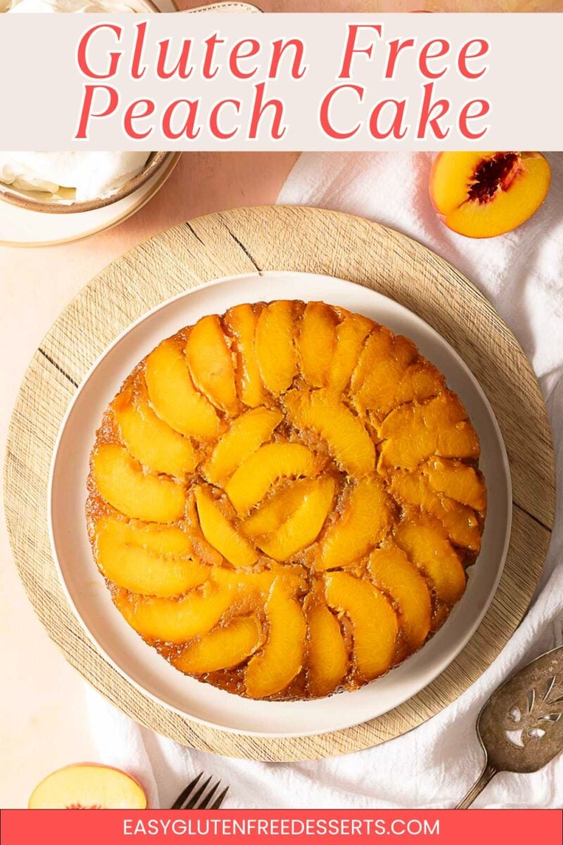 A round gluten free peach upside down cake topped with sliced peaches sits on a white plate, placed on a wooden mat. Sliced peaches and a bowl of whipped cream are nearby.