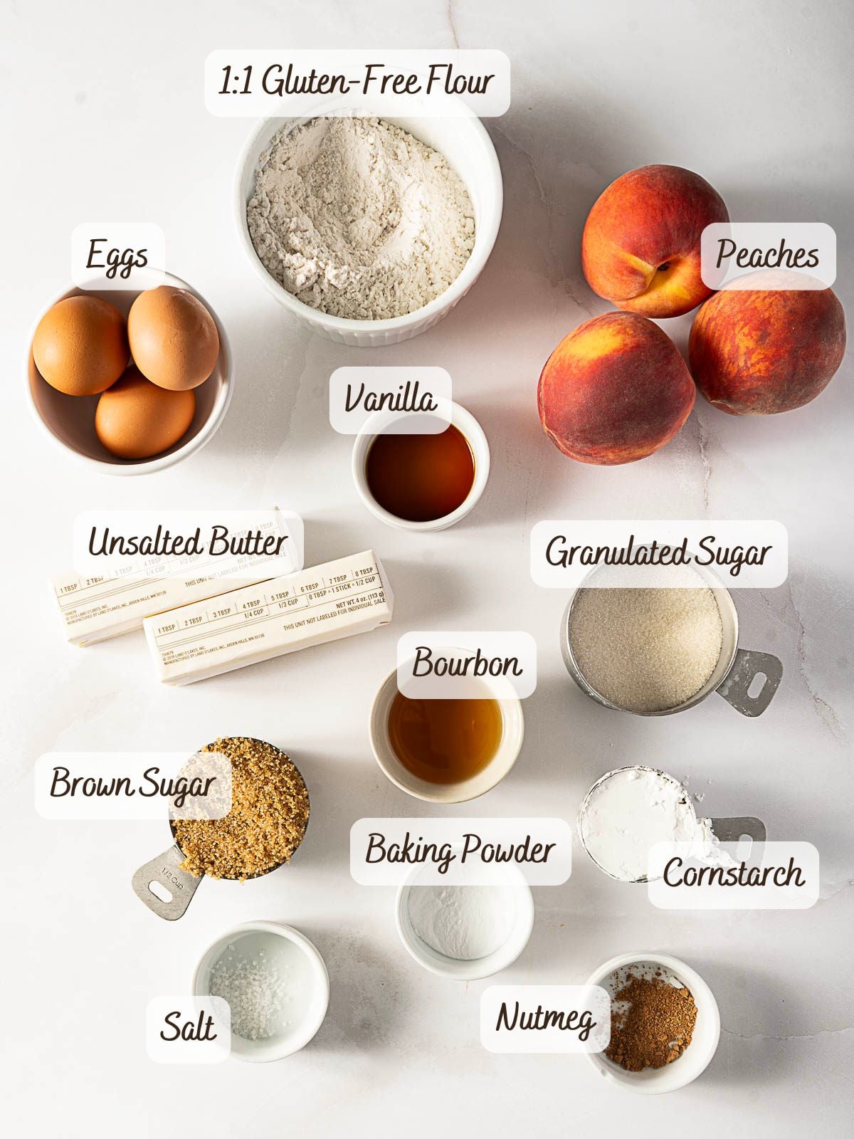 Ingredients for a peach upside down cake are arranged on a white surface, including flour, eggs, peaches, vanilla, butter, sugars, bourbon, baking powder, cornstarch, salt, and nutmeg.