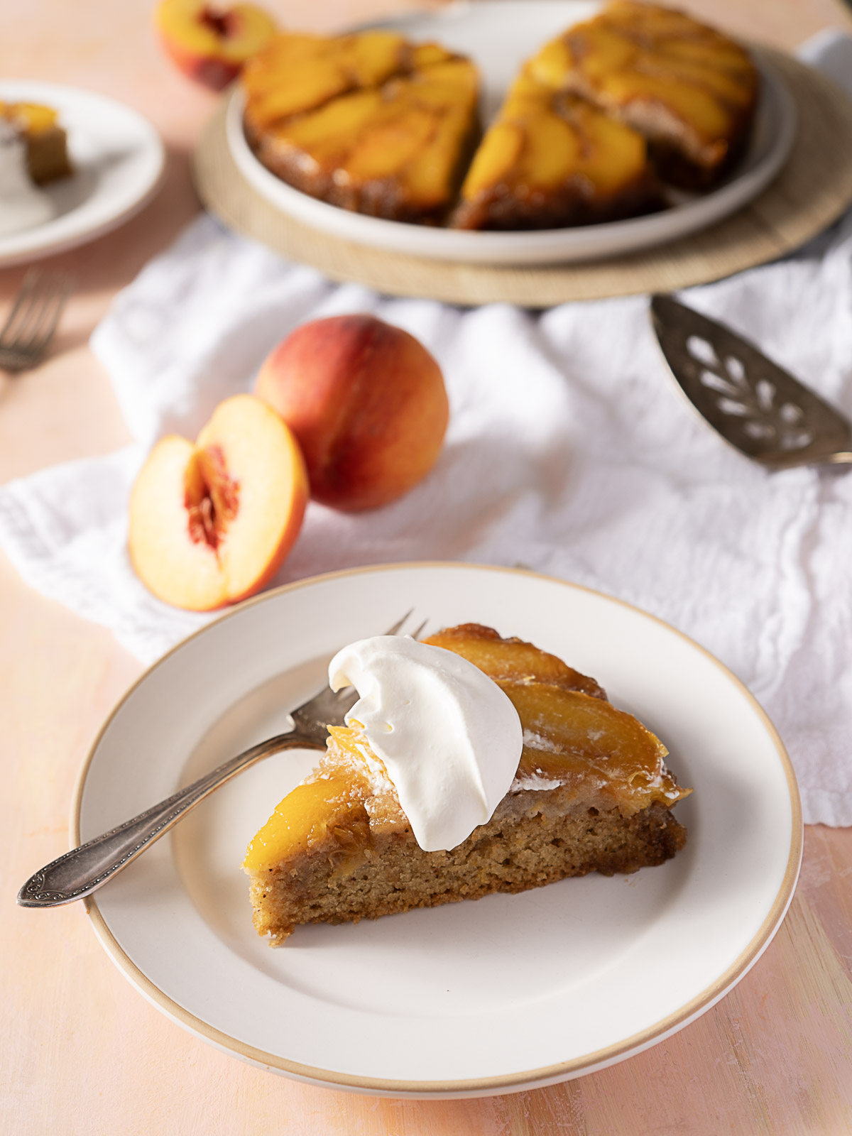 A slice of peach upside-down cake with whipped cream on a plate, a fork, fresh peach halves, and the rest of the cake in the background.