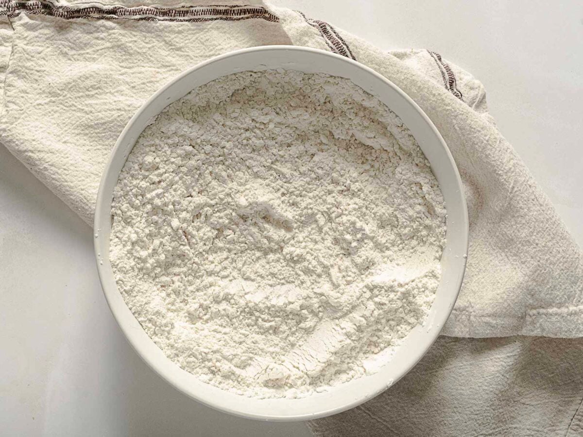 A white bowl filled with flour sits on a cream-colored textured cloth on a white surface.