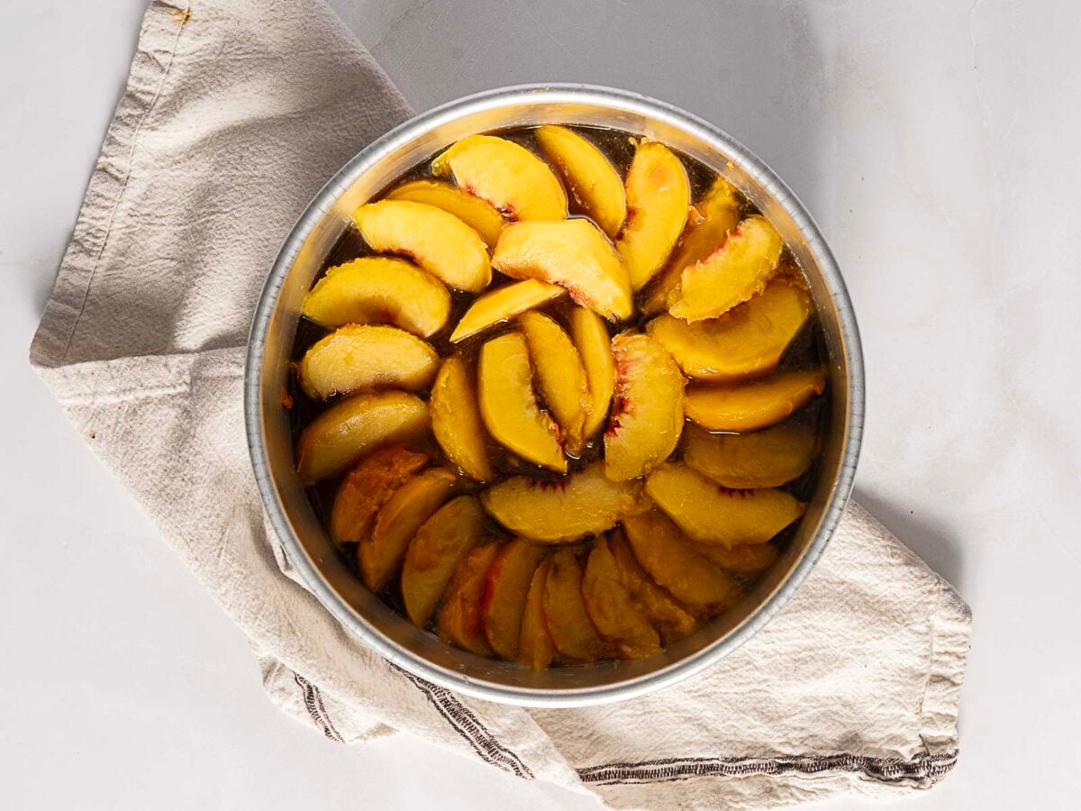 A metal bowl filled with sliced peaches soaking in liquid, placed on a folded beige kitchen towel on a light-colored surface.