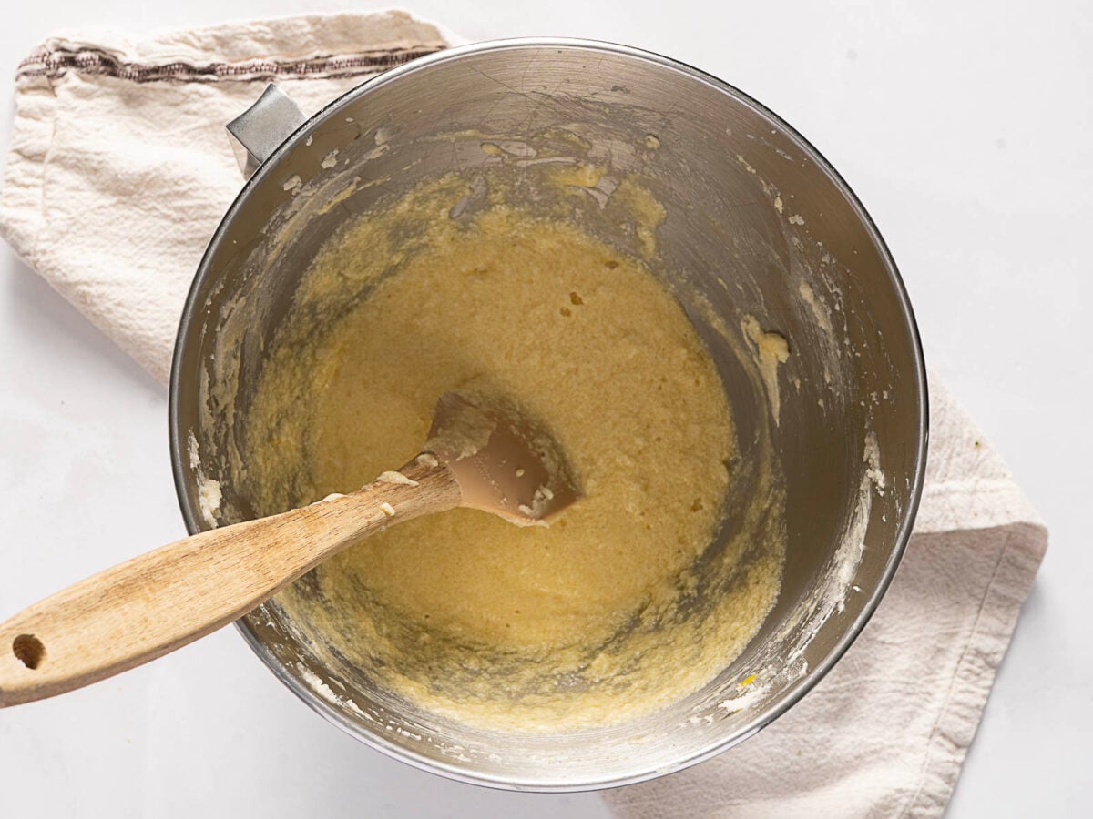 A mixing bowl filled with yellow cake batter and a wooden spatula, placed on a beige kitchen towel on a white surface.