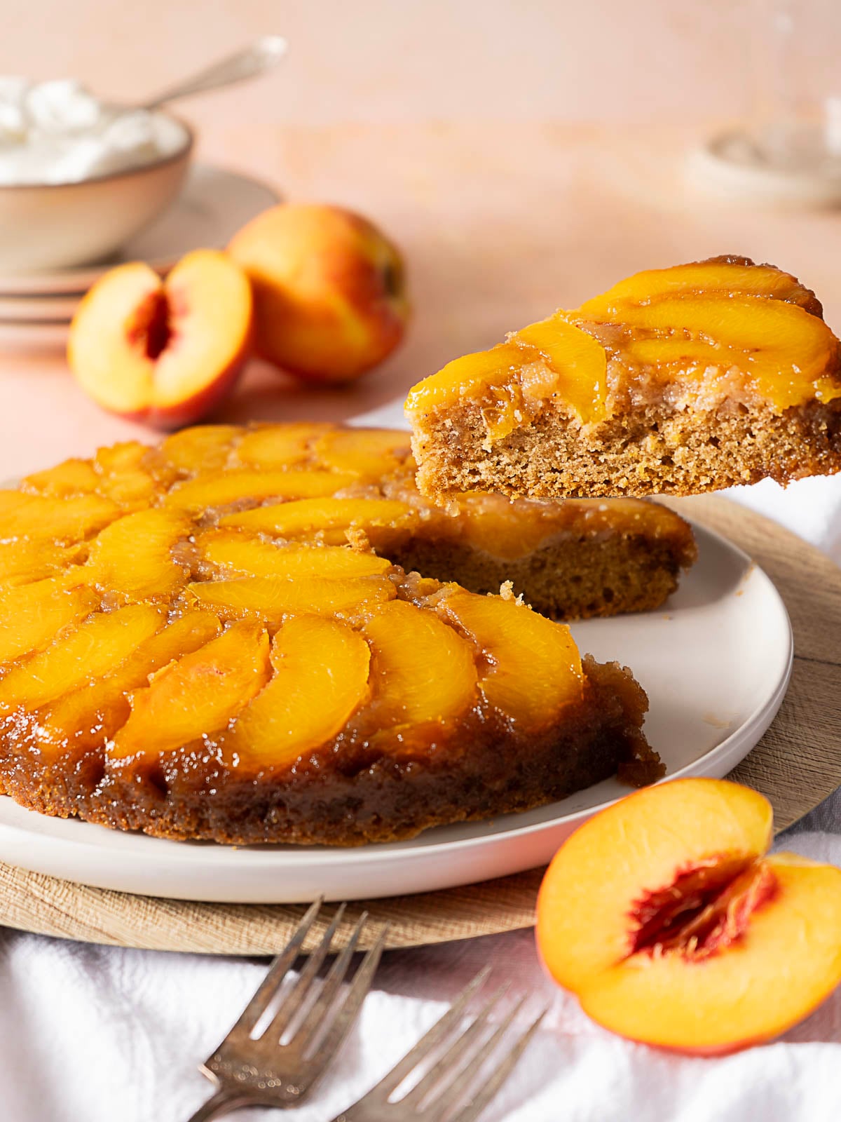 A peach upside-down cake on a white plate, with one slice being lifted; fresh peach halves and forks are nearby.