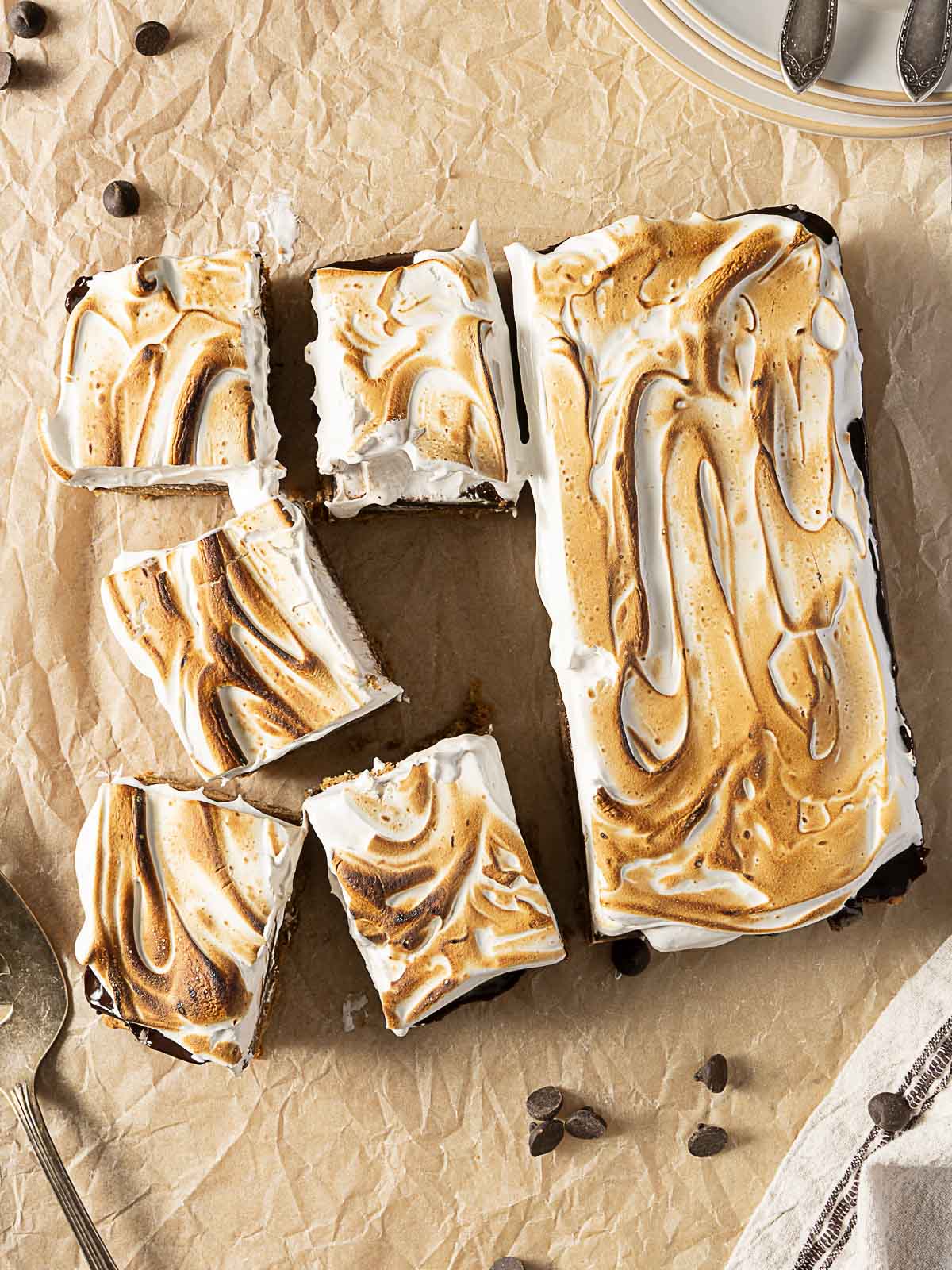 A rectangular cake with toasted swirled meringue topping sits on parchment paper, partially sliced into square pieces. Chocolate chips are scattered around.