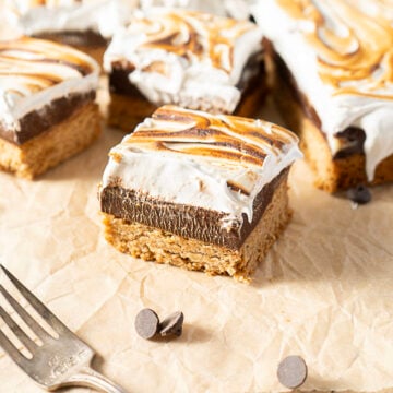 A square dessert bar with a cookie base, chocolate layer, and toasted marshmallow topping sits on parchment paper near a fork and chocolate chips.