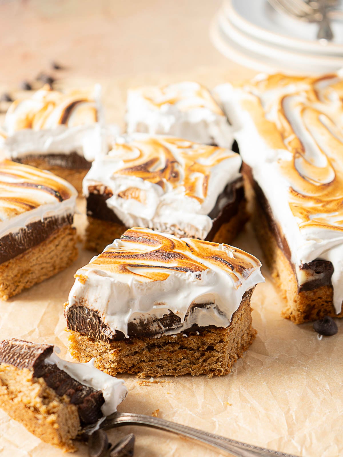 Squares of layered dessert bars with a graham base, chocolate middle, and toasted meringue topping are arranged on parchment paper, with one piece being lifted by a fork.