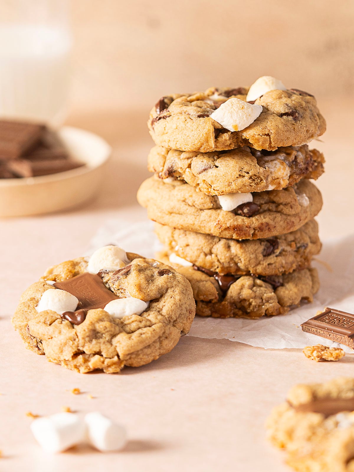 A stack of chocolate chip cookies with marshmallows and chocolate pieces, with one cookie in front and a glass of milk and chocolate pieces in the background.
