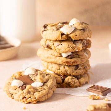 A stack of chocolate chip cookies with marshmallows sits on parchment paper next to a cookie with a chocolate piece on top and a chocolate square.