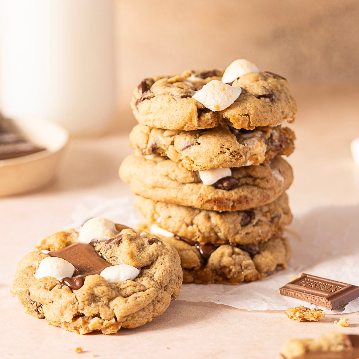 A stack of chocolate chip cookies with marshmallows sits on parchment paper next to a cookie with a chocolate piece on top and a chocolate square.