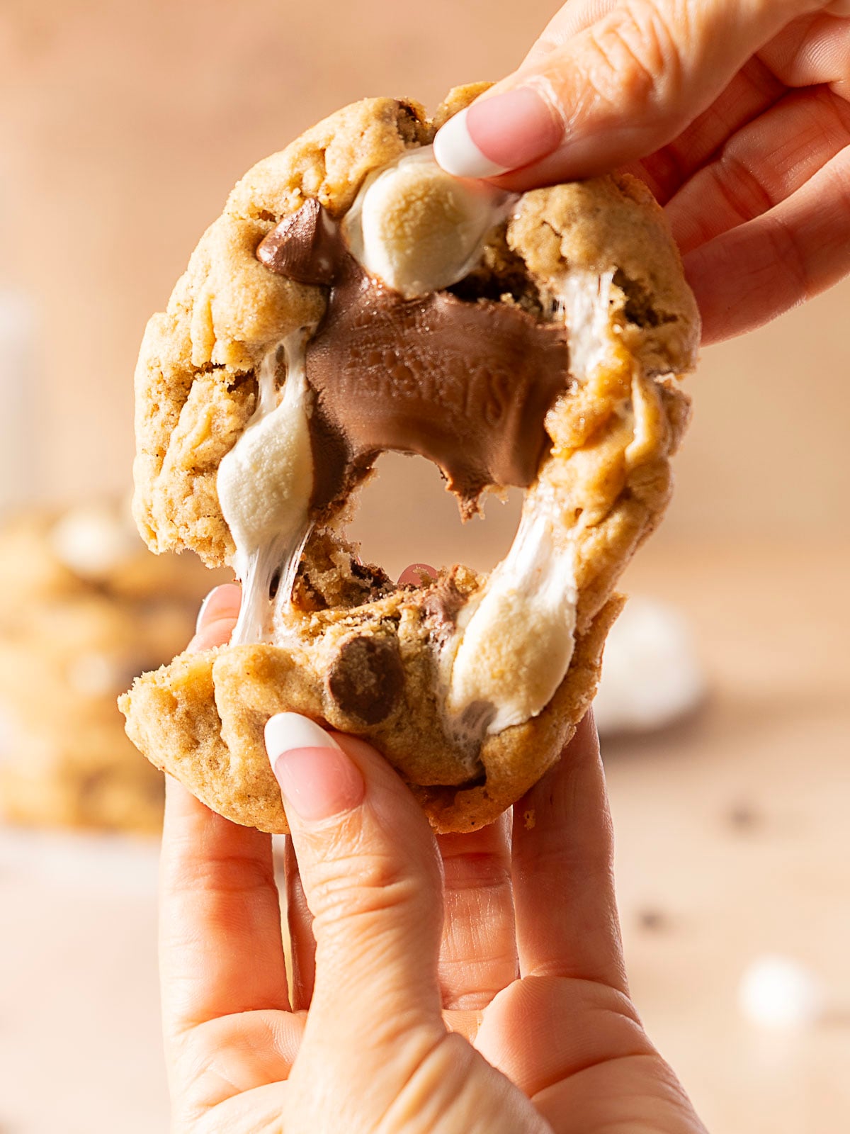 Two hands pull apart a thick chocolate chip cookie with melted chocolate and marshmallow filling stretching between the halves.