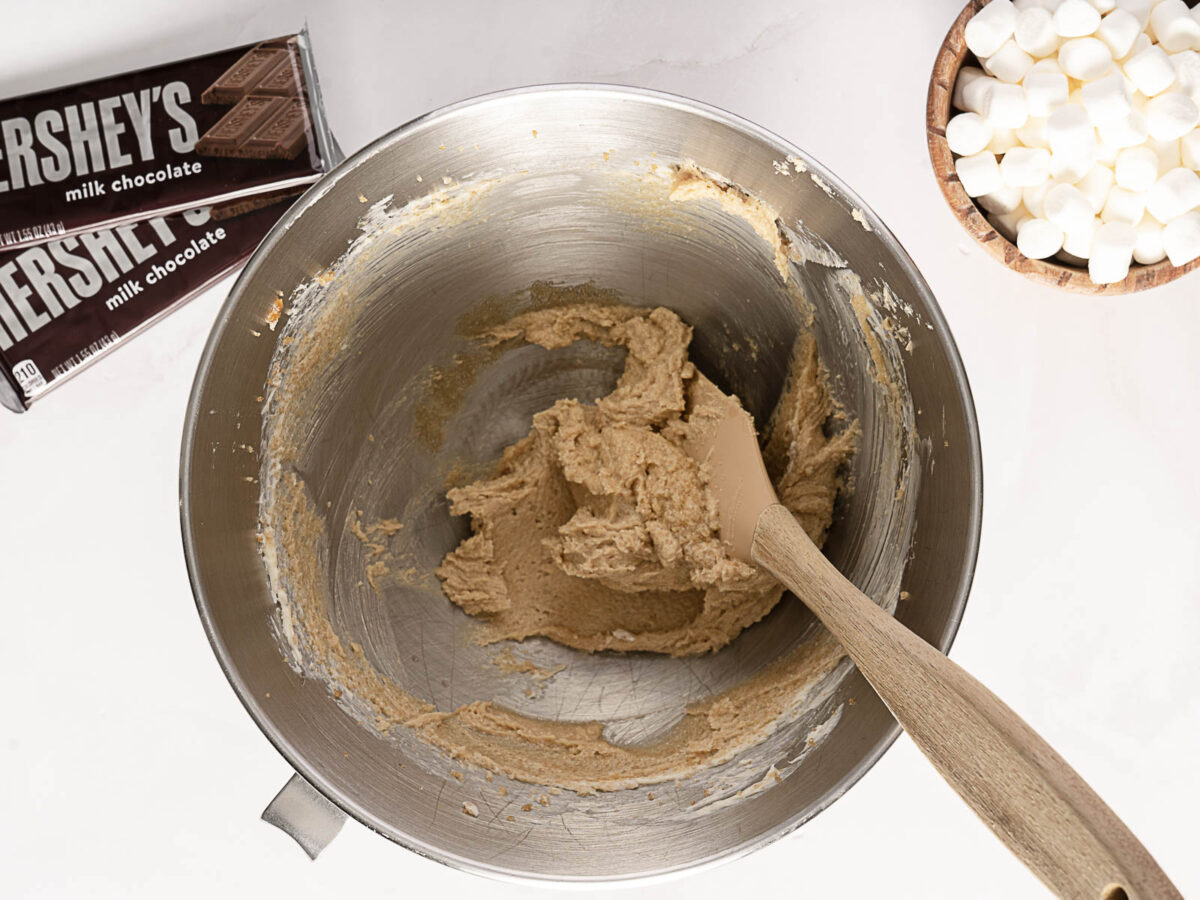 A metal mixing bowl with cookie dough, a spatula, a bowl of mini marshmallows, and two Hershey's milk chocolate bars on a white countertop.