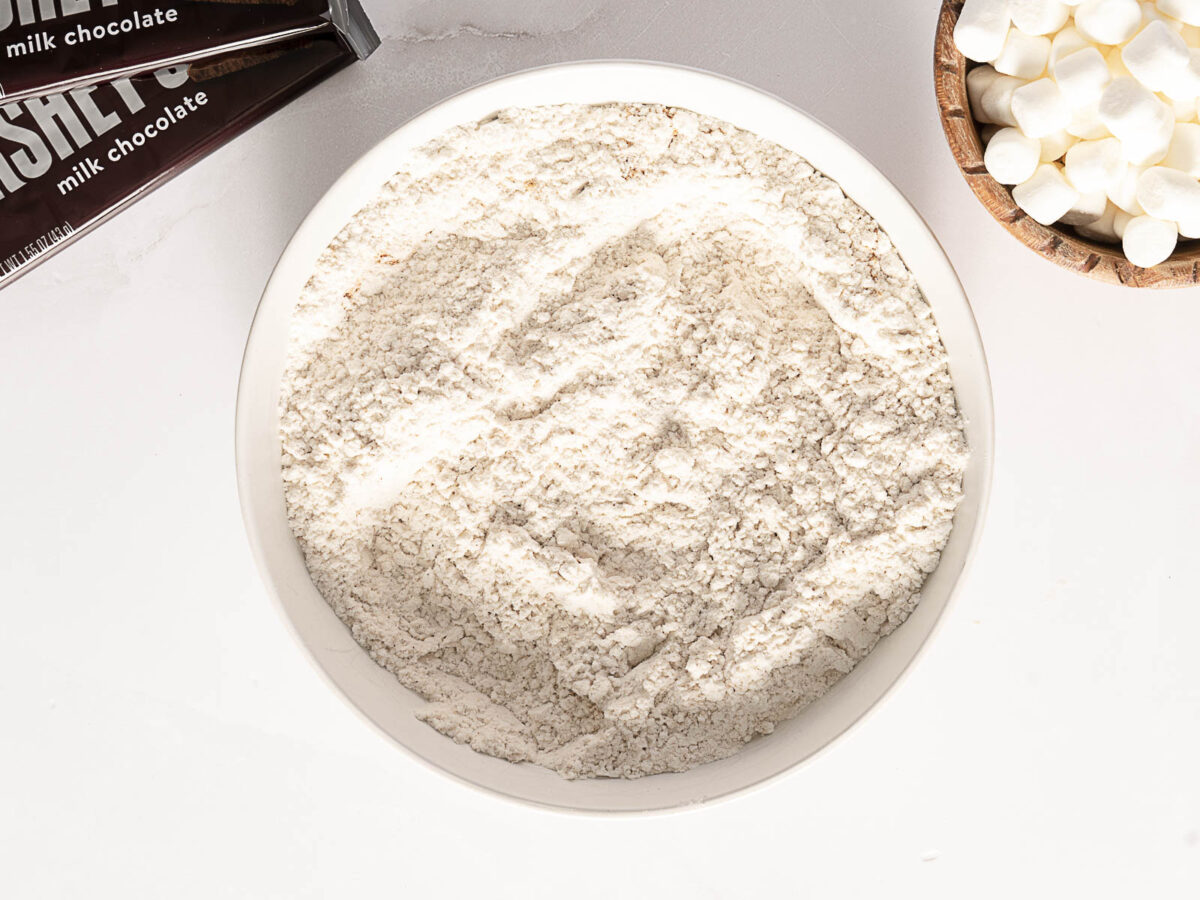 A bowl of flour is centered on a white surface, with a chocolate bar and a small bowl of marshmallows nearby.