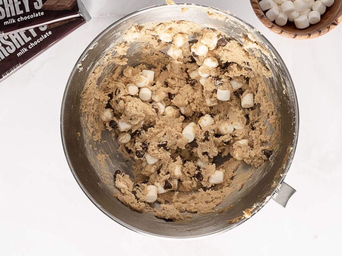 A metal mixing bowl with cookie dough containing chocolate chips and mini marshmallows, with chocolate bars and a bowl of marshmallows nearby.
