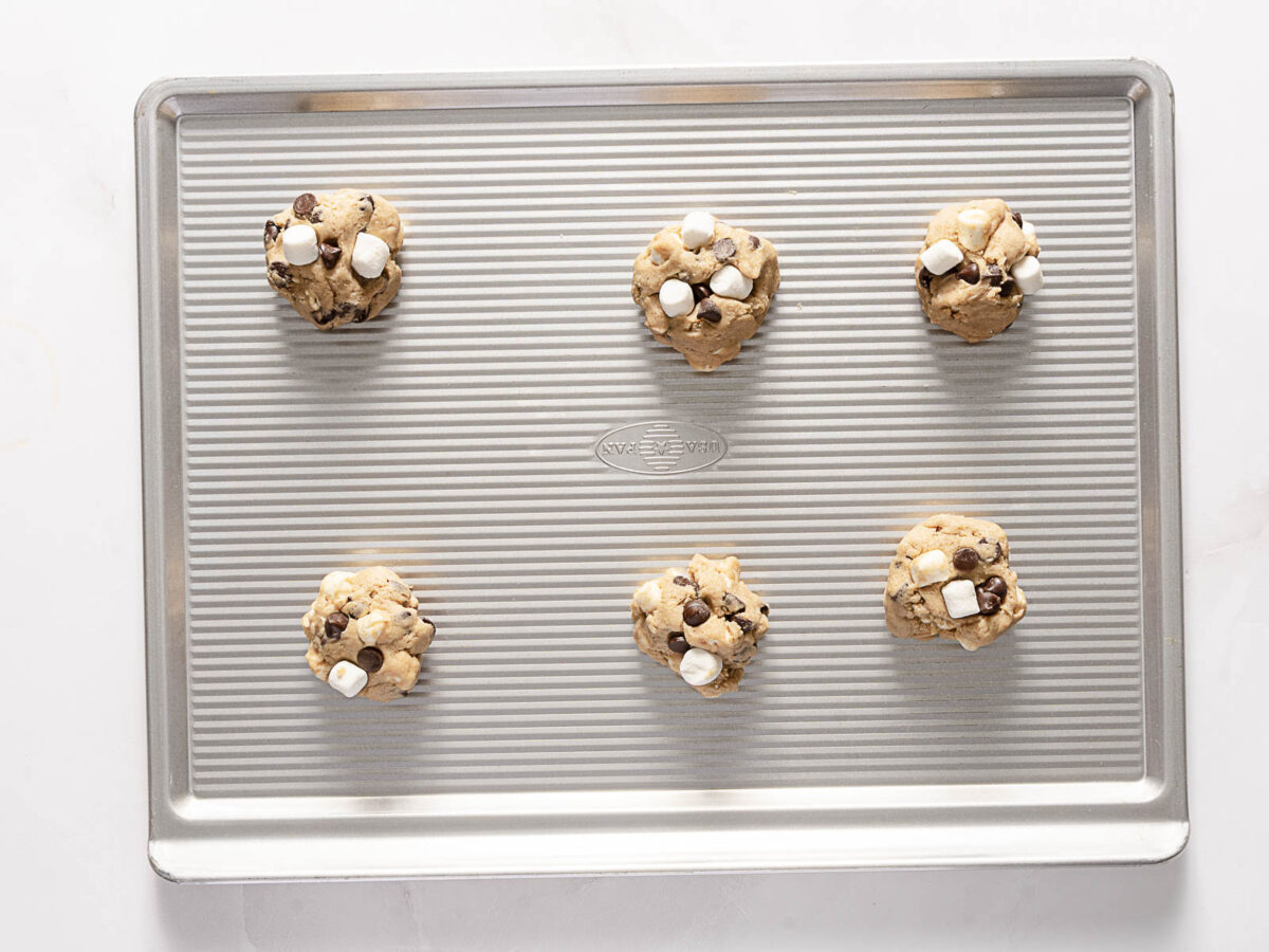 Six unbaked cookie dough balls with chocolate chips and marshmallows arranged on a ridged baking sheet.
