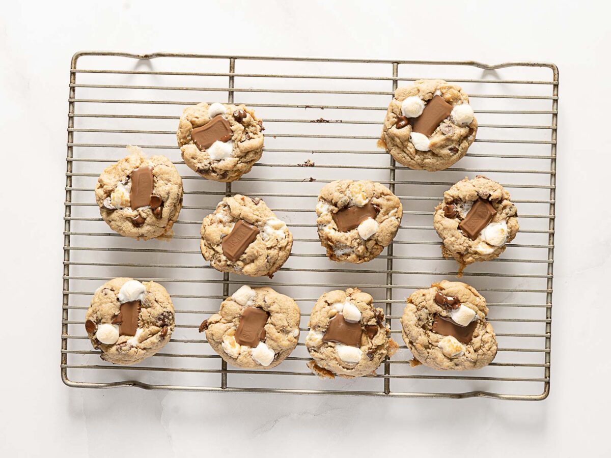 Ten cookies with chocolate pieces and marshmallows are arranged on a metal cooling rack against a white background.