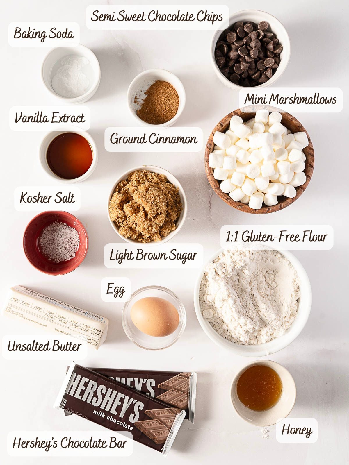 Top-down view of labeled baking ingredients in bowls and cups, including chocolate chips, marshmallows, flour, sugar, butter, vanilla, cinnamon, egg, salt, baking soda, honey, and chocolate bar-perfect for making delicious Gluten Free S'Mores Cookies.
