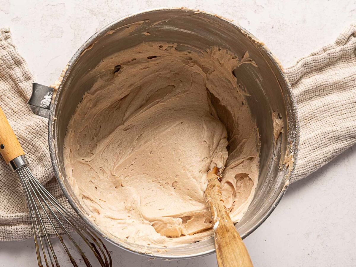 Metal bowl with light brown whipped mixture, a wooden spoon inside, a whisk nearby, and a beige kitchen towel underneath on a white surface.