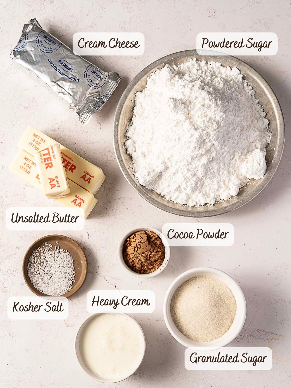 Ingredients for Chocolate Caramel Frosting are arranged on a countertop, including cream cheese, powdered sugar, unsalted butter, kosher salt, cocoa powder, heavy cream, and granulated sugar—each clearly labeled with text.
