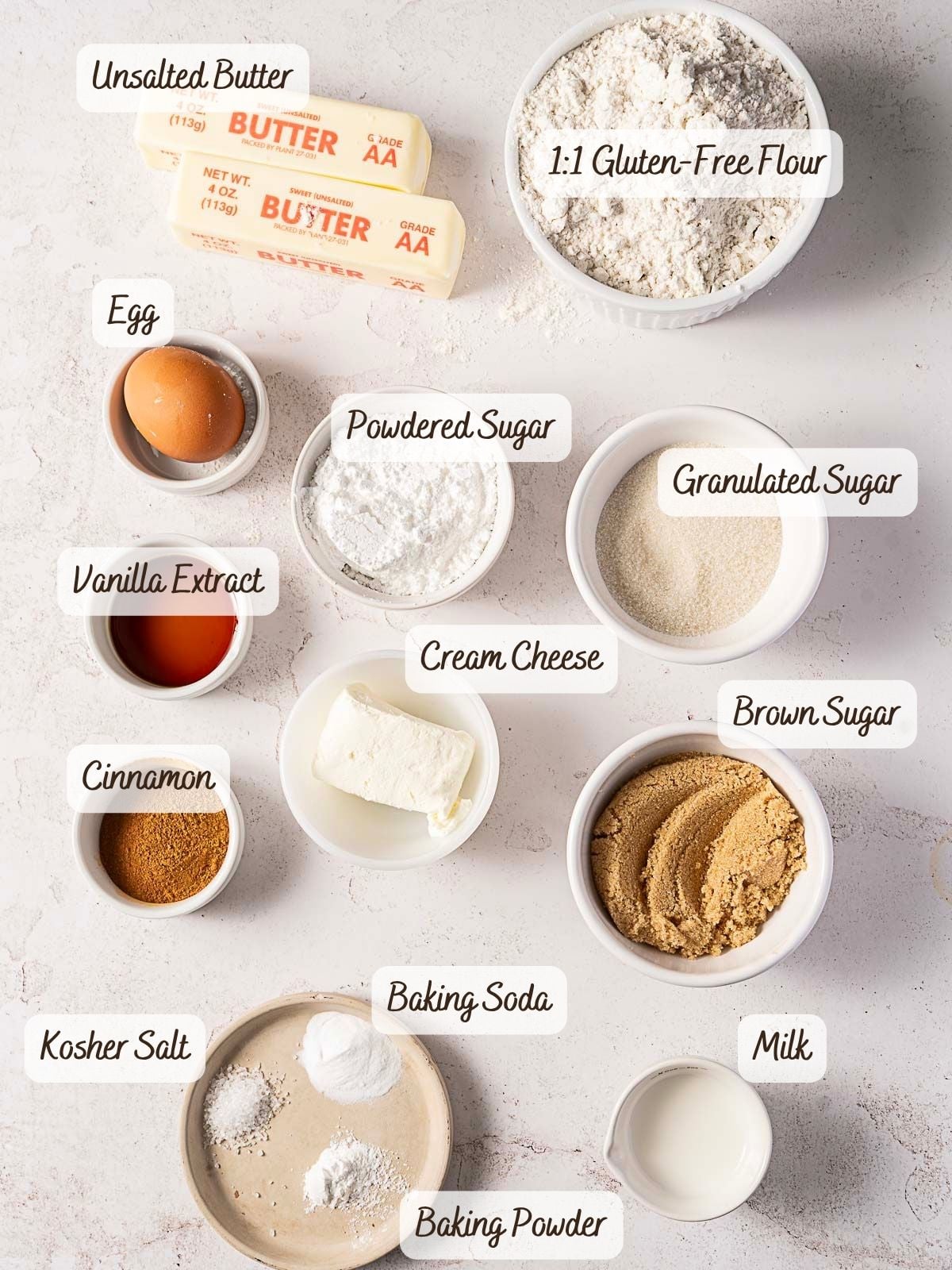 Various baking ingredients for Gluten-Free Cinnamon Roll Cookies—including unsalted butter, gluten-free flour, sugars, egg, vanilla, cream cheese, cinnamon, salt, baking soda, baking powder, and milk—arranged on a light surface.