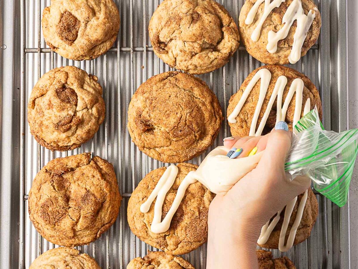 A hand pipes white icing onto cinnamon cookies arranged on a metal cooling rack.