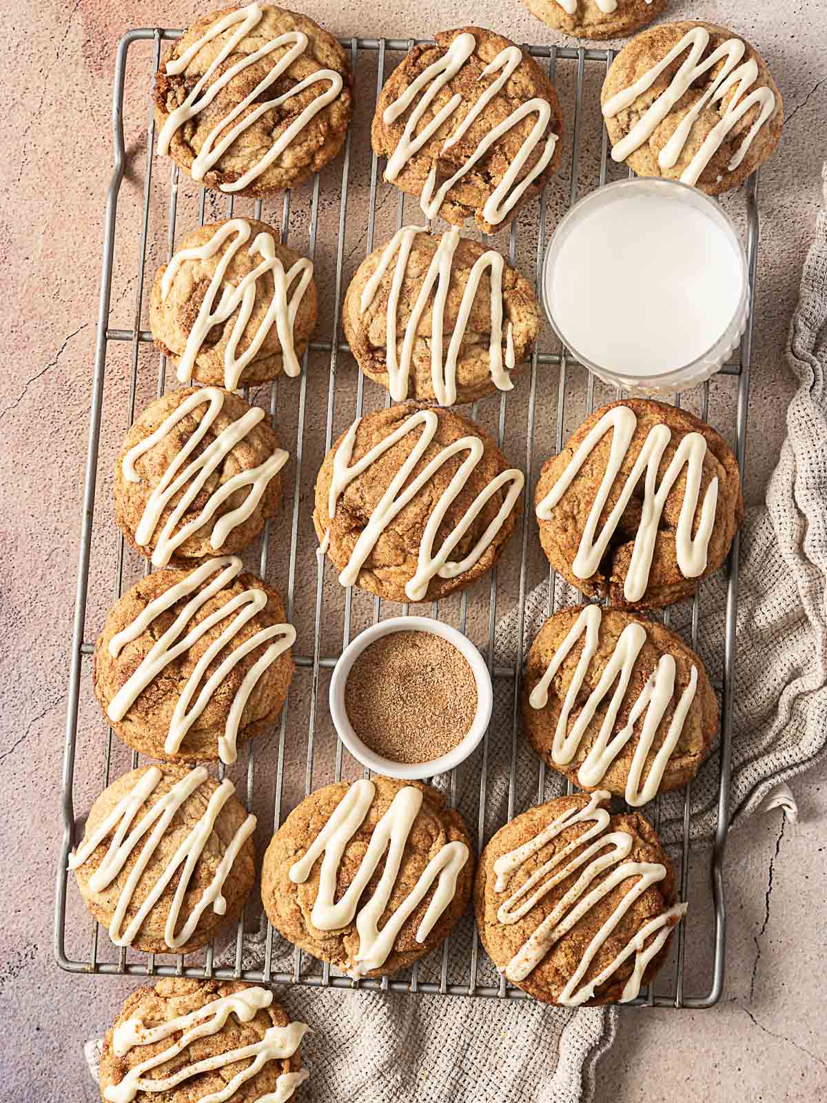 Cookies with white icing drizzle arranged on a cooling rack, accompanied by a small bowl of cinnamon sugar and a cup of milk on a textured surface.
