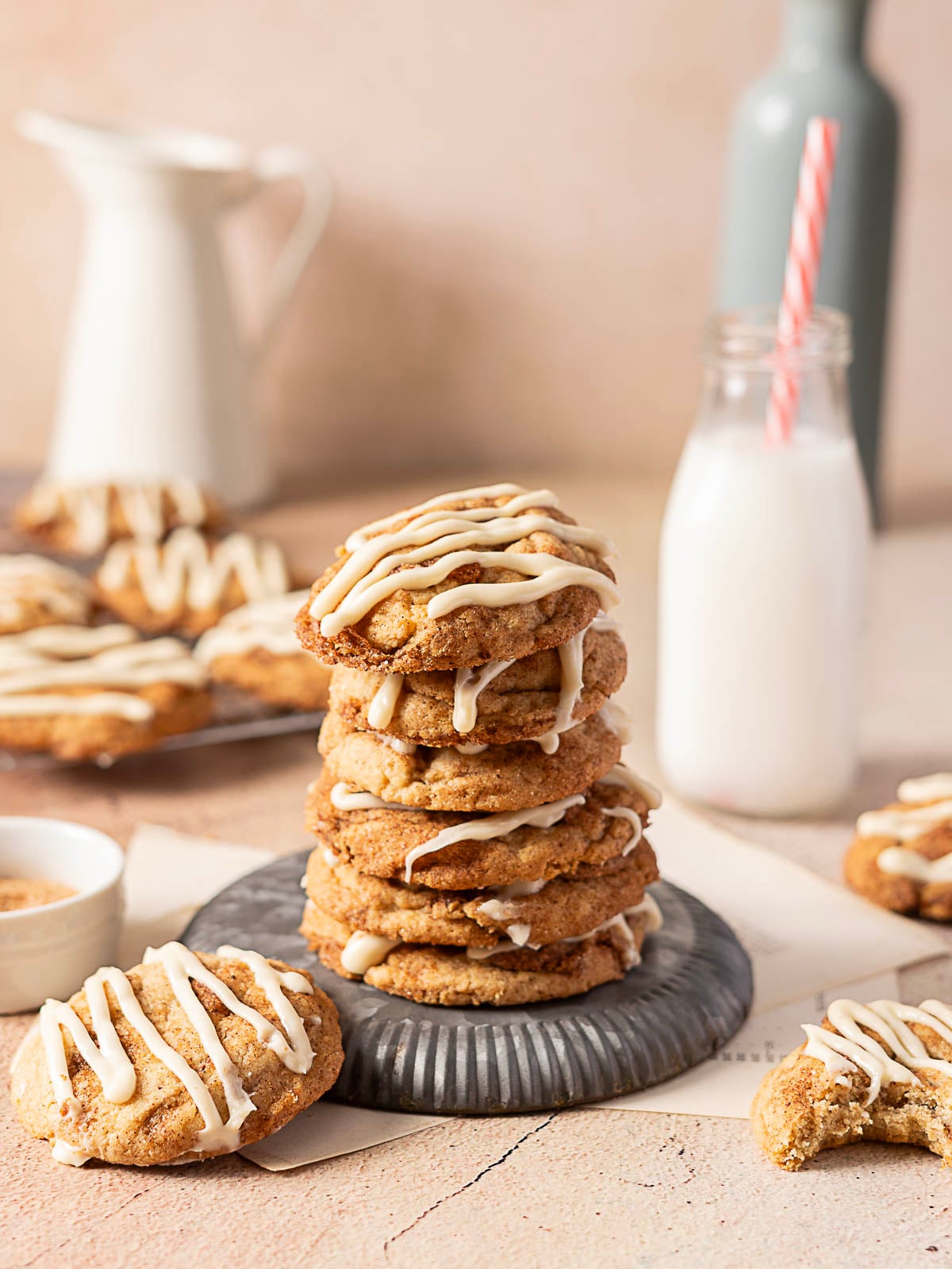 A stack of glazed cookies is on a plate, surrounded by more cookies, a bottle of milk with a straw, and a small bowl of sugar on a light-colored surface.