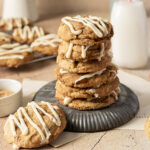 A stack of six cookies with white icing drizzle sits on a metal plate, surrounded by more cookies and a glass of milk in the background.