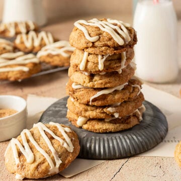 A stack of six cookies with white icing drizzle sits on a metal plate, surrounded by more cookies and a glass of milk in the background.