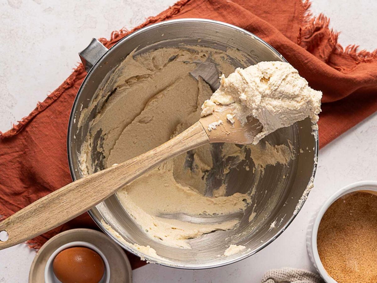 A metal mixing bowl with creamy batter and a wooden spatula, next to a brown egg in a dish and a bowl of cinnamon sugar, on a rust-colored cloth.