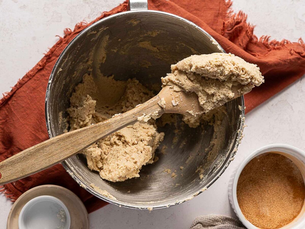 A wooden spoon rests on a mixing bowl filled with cookie dough, placed on a rust-colored cloth, with a small bowl of brown sugar nearby.