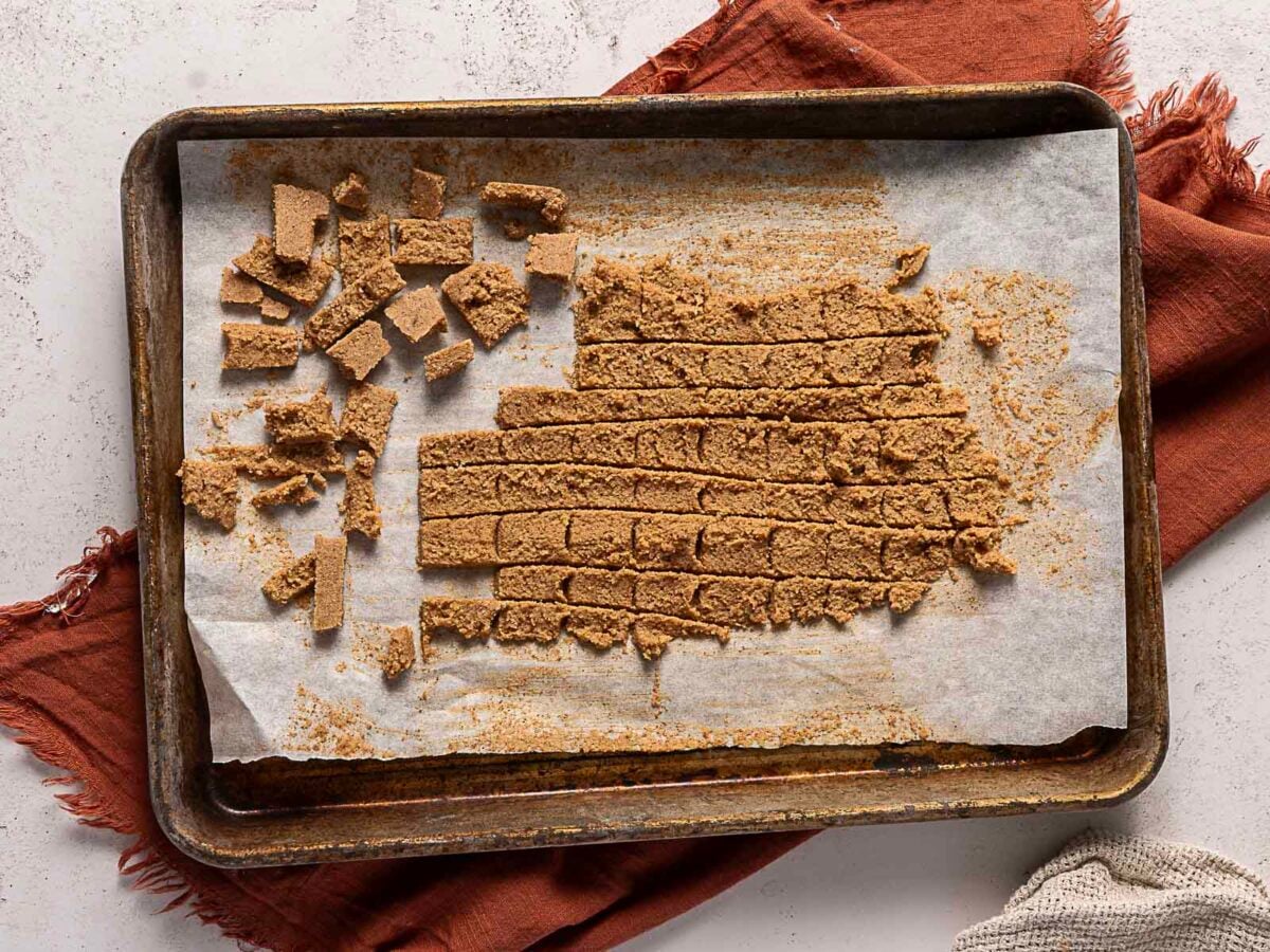 A baking sheet with parchment paper holds cut and uncut pieces of brown crumbly dough, some pieces separated on the left side. A brown cloth is partially visible underneath the tray.