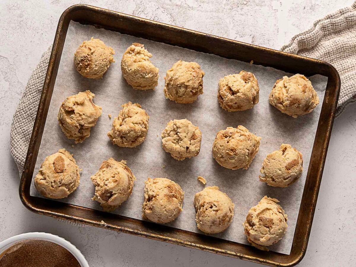 A baking sheet lined with parchment paper holds 16 scoops of raw cookie dough, spaced evenly apart, ready to be baked. A beige kitchen towel and a bowl are nearby.