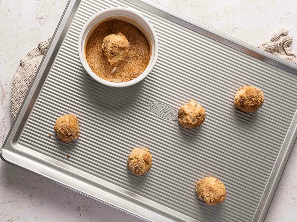 Five unbaked cookie dough balls on a metal baking sheet; one is in a small bowl of cinnamon sugar, ready to be coated.