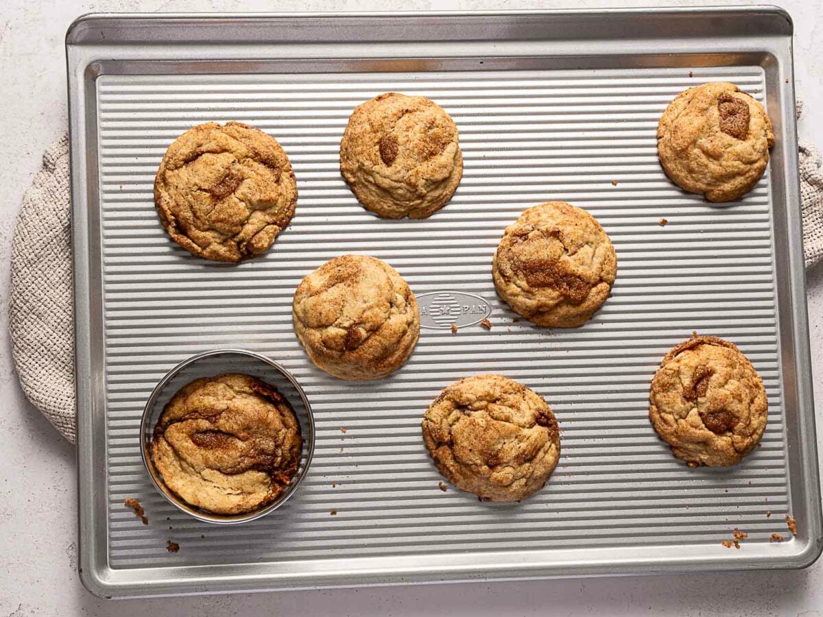 Seven cookies are on a ridged baking sheet, with one cookie partially cut by a round metal biscuit cutter. A textured cloth is visible underneath the sheet.