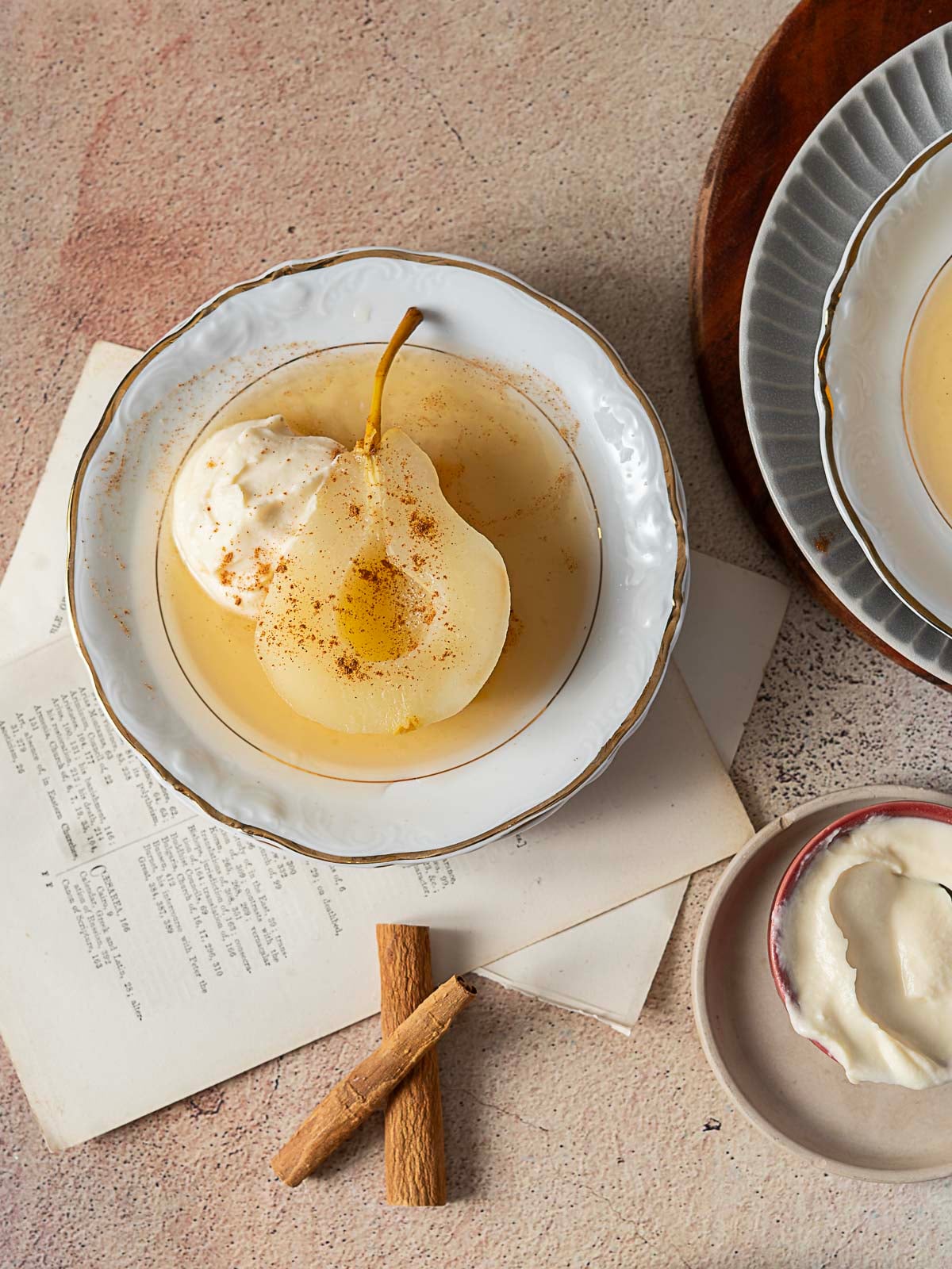 A poached pear with whipped cream and a sprinkle of cinnamon on a white plate, next to cinnamon sticks, a bowl of cream, and an open recipe book.
