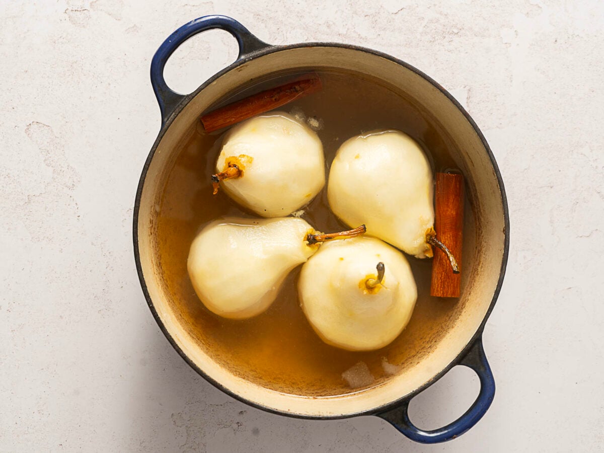 Four peeled pears and two cinnamon sticks simmer in a pot of liquid.