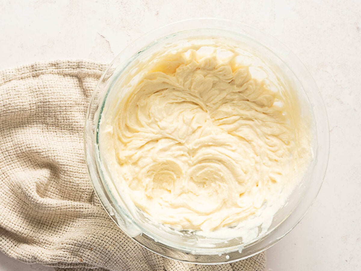A glass bowl filled with creamy cake batter sits on a textured beige cloth atop a light-colored surface.
