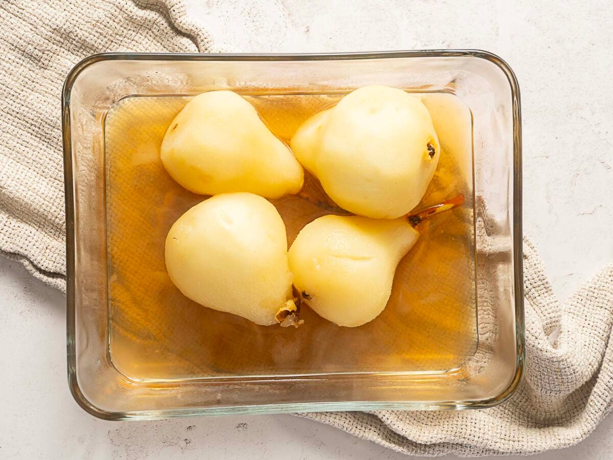 Four peeled pears are placed in a glass dish with liquid, set on top of a textured beige cloth on a light-colored surface.