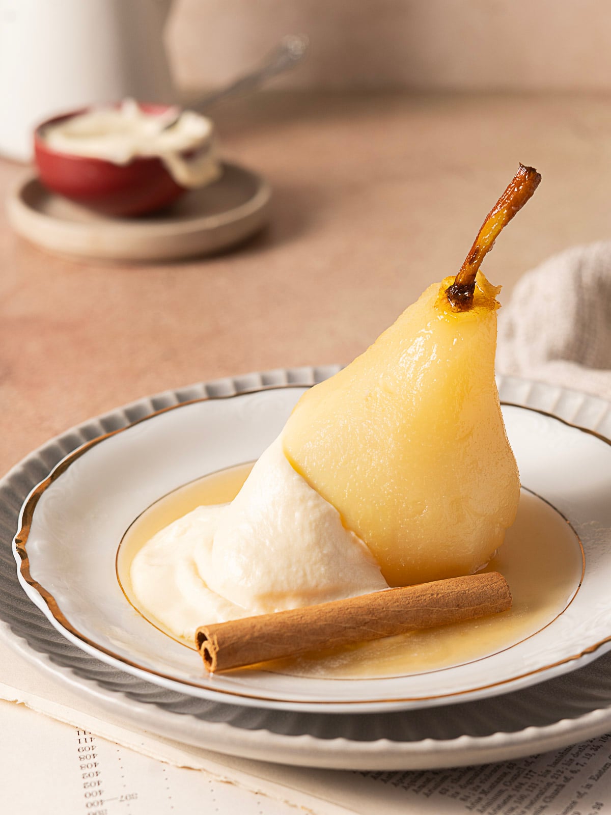 A poached pear is served on a white plate with whipped cream, syrup, and a cinnamon stick. A small bowl with a spoon is in the blurred background.
