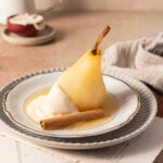 A poached pear served with whipped cream and a cinnamon stick on a white plate, placed on a stacked plate setting.
