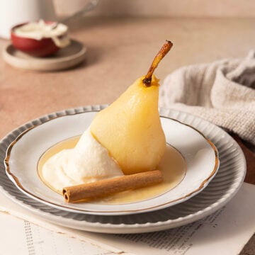 A poached pear served with whipped cream and a cinnamon stick on a white plate, placed on a stacked plate setting.