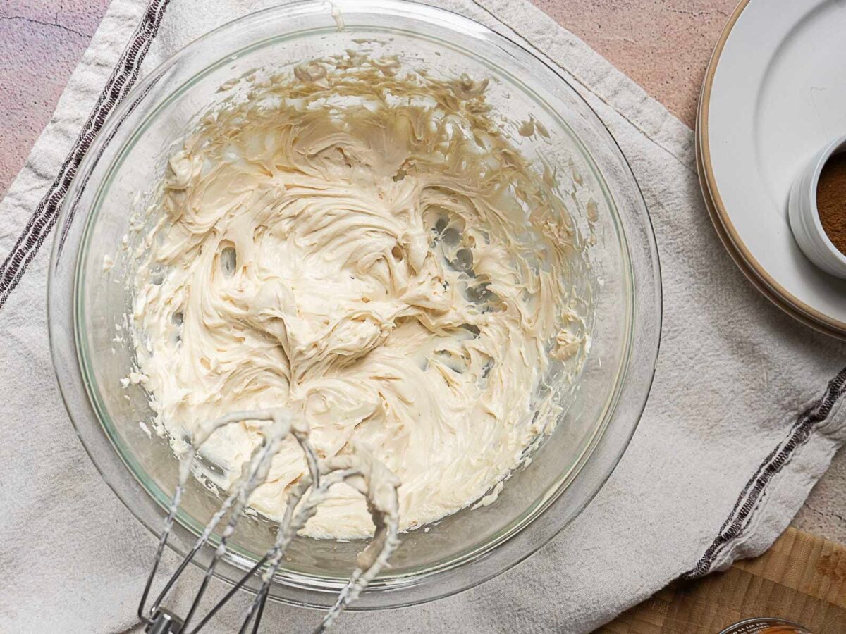A glass bowl with whipped cream cheese mixture and a hand mixer on a white towel, with plates and a small bowl nearby.