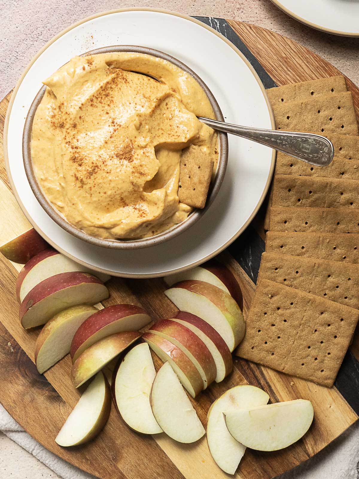 Bowl of creamy dip with a spoon, served with rectangular crackers and sliced red and green apples on a wooden board.
