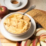 Bowl of creamy dip with graham crackers and apple slices on a wooden board, with an apple and small dish in the background.
